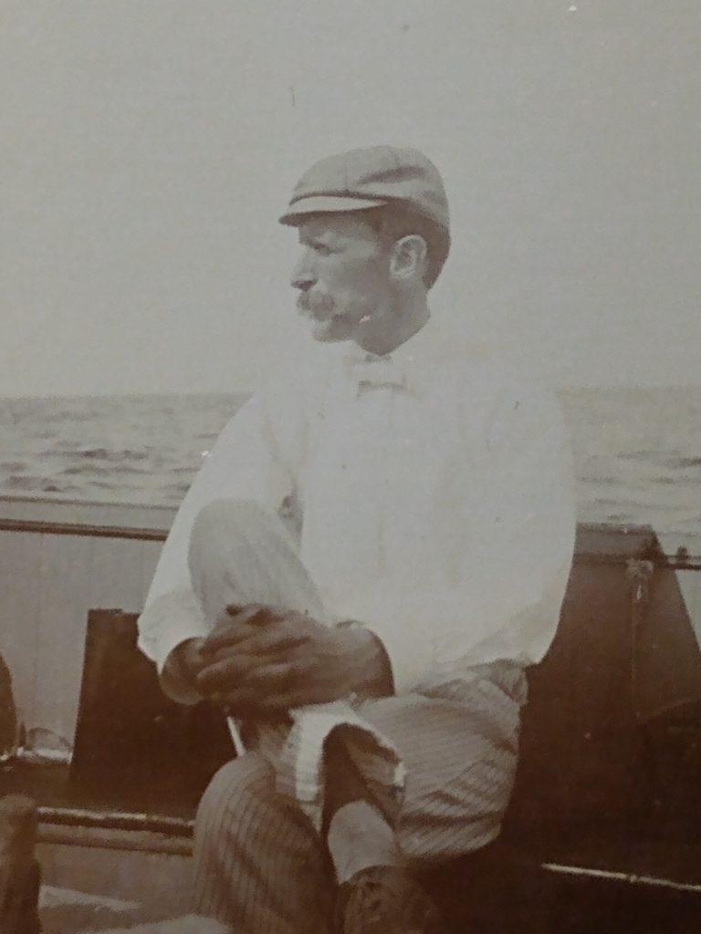 Black and white photograph of a white man with short dark hair and a mustache sitting inside a boat with the water behind him. His left leg is crossed over his right leg, and his hands are cupping his left shin. He wears a cloth cap, a long-sleeved white shirt, a white bowtie, and striped pants with cuffs. He looks off to his left.